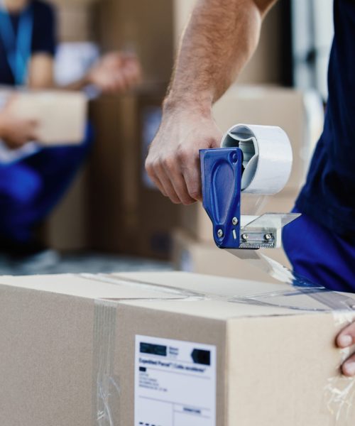 Close-up of delivery man closing carboard box with a tape while preparing packages for shipment.